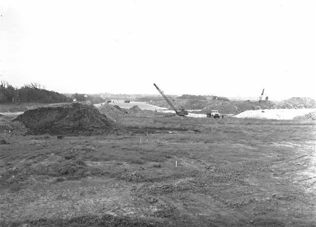 Grondwerken door N.V. B. Smallegange voor de aanleg van de aansluiting van de geplande provinciale weg nr. 4 (tussen Noordwijk en Oegstgeest) in De Klei bij De Krom. Links het dak van café De Viersprong. Rechts, boven de witte vrachtwagen, het dak van een gebouw van het magazijncomplex van Defensie, De Krom. Op de achtergrond de bebouwing van Katwijk.