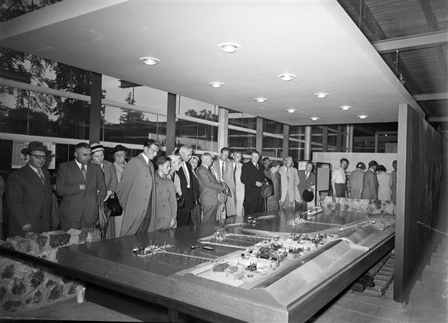 Leden van de Provinciale Staten bekijken de stand van de provincie Zuid-Holland, op de Nationale Energie Manifestatie 1955 (E55). De maquette geeft de verschillende stadia weer van de dijkaanleg bij de Hoek van Sint Jacob, op Goeree-Overflakkee, na de watersnoodramp van 1953.