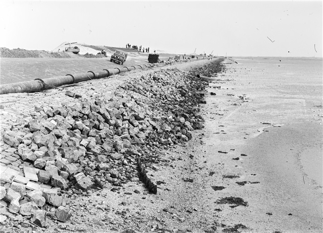 Dijkherstel bij Oude-Tonge na de watersnoodramp van 1 februari 1953. Het nieuwe dijklichaam van opgespoten zand wordt bekleed met asfalt. De glooiing onderaan de dijk wordt afgewerkt met stenen.
