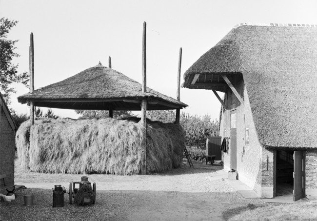Gedeelte van een boerderij met een meerroedige hooiberg. Op de voorgrond een melkbussenkar en rechts aan de muur een zaaijuk.