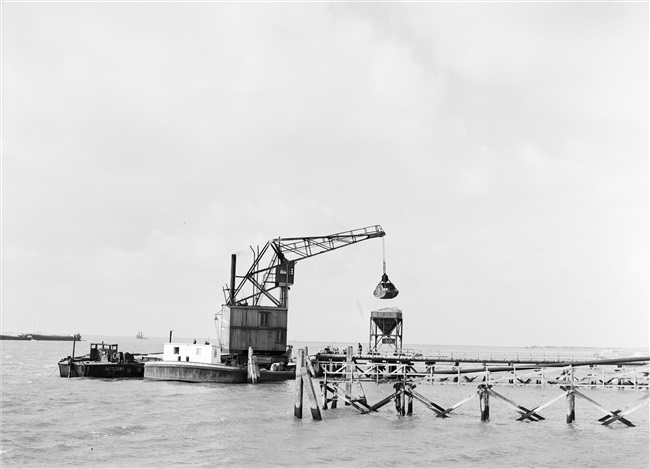 Dijkherstel op Goeree-Overflakkee, na de watersnoodramp van 1 februari 1953. Uit een binnenvaartschip wordt zand overgeladen in een tijdelijke opslag, voor de productie van zandasfalt. In de polder Galathee (Ooltgensplaat) staat o.a. een zandasfaltfabriek. Met het geproduceerde asfalt worden de herstelde dijken bekleed.