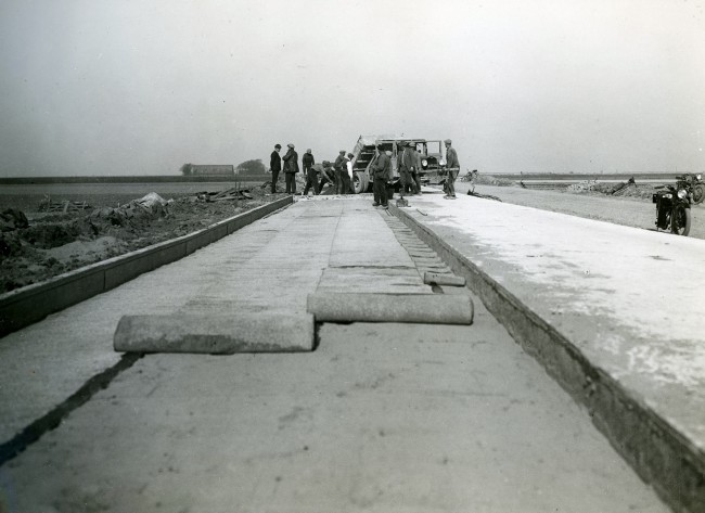 De aanleg van een weg door de Provinciale Waterstaat in Zuid-Holland. Op een zandlaag wordt asfaltpapier aangebracht. De zandauto draagt het kenteken H85353 dat in 1930 is uitgegeven. De foto is waarschijnlijk gemaakt tussen 1930 en 1950.