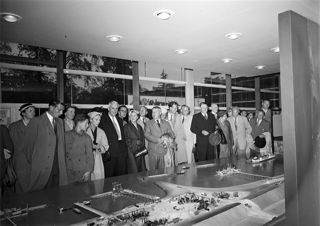 Leden van de Provinciale Staten bekijken de stand van de provincie Zuid-Holland, op de Nationale Energie Manifestatie 1955 (E55). De maquette geeft de verschillende stadia weer van de dijkaanleg bij de Hoek van Sint Jacob, op Goeree-Overflakkee, na de watersnoodramp van 1953.