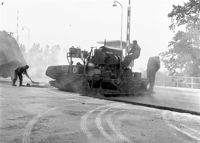 Een asfaltwerker en een asfaltmachinist brengen de asfalt wegverharding aan op de eerste St. Sebastiaansbrug.