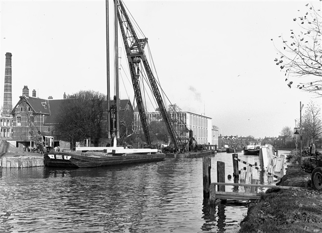 Bouw van de eerste St. Sebastiaansbrug over het Rijn-Schiekanaal. Een drijvende bok wordt gebruikt bij het heien van de palen. Op de achtergrond het voormalige microbiologielaboratorium van de Polytechnische School.