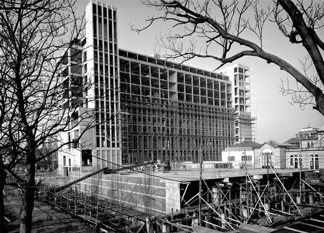 Het betonskelet van de hoogbouw van het nieuwe provinciehuis; aanzicht van de achtergevel met twee trappenhuizen. Op de voorgrond de bouw van de laagbouw, o.a. bestemd voor het college van Gedeputeerde Staten. Rechts het Moorse Paleis van de voormalige Haagsche Dierentuin.