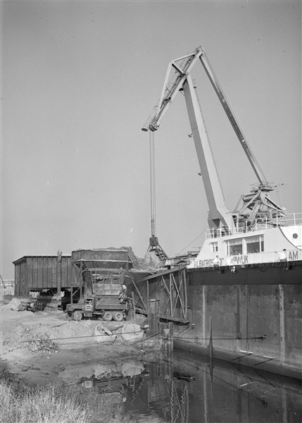 Aanleg van een weg op zandpalen. Met een grijper wordt het per schip aangevoerde zand in een silo gestort en vervolgens met zandauto's afgevoerd.