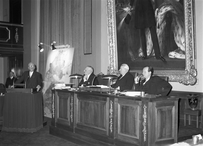 De Provinciale Staten van Zuid-Holland vergaderen in het gebouw van de Eerste Kamer. Onder het schilderij in het midden de heer A.C.A. Deerenberg en links van hem griffier F.A. Helmstrijd.