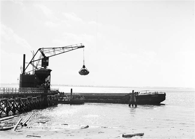 Dijkherstel op Goeree-Overflakkee, na de watersnoodramp van 1 februari 1953. Uit een binnenvaartschip wordt zand overgeladen in een tijdelijke opslag, voor de productie van zandasfalt. In de polder Galathee (Ooltgensplaat) staat o.a. een zandasfaltfabriek. Met het geproduceerde asfalt worden de herstelde dijken bekleed.