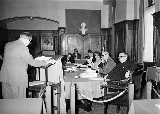 Een vergadering van de Gedeputeerde Staten aan het Korte Voorhout 1. Aan tafel zitten van rechts naar links: gedeputeerde mr. H. van Riel, griffier mr. F.A. Helmstrijd, gedeputeerden dr. J.J.R. Schmal, mevrouw mr. Chr. A. de Ruyter-de Zeeuw en J.P. van Praag.