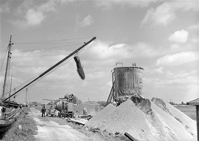 Aanleg van de weg op palen tussen Boskoop en Alphen aan den Rijn, door de Provinciale Waterstaat. Een baggerschip stort grint, wat wordt gebruikt voor de aanmaak van beton.