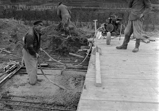 Aanleg van de weg op palen tussen Boskoop en Alphen aan den Rijn. Met een pneumatische vibrator wordt beton aangetrild, dat vanaf een verrijdbare vlonder (rechts) in de wapening voor het wegdek is gestort.