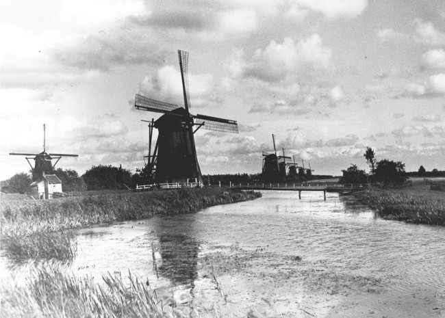 De molens van Kinderdijk in de polders van de Alblasserwaard. Op de voorgrond molen nr. 5 van de Nederwaard en van links naar rechts de molens 3 t/m 8 van de Overwaard. Links op de voorgrond een bakhuisje, waarin men vroeger brood en koek bakte.