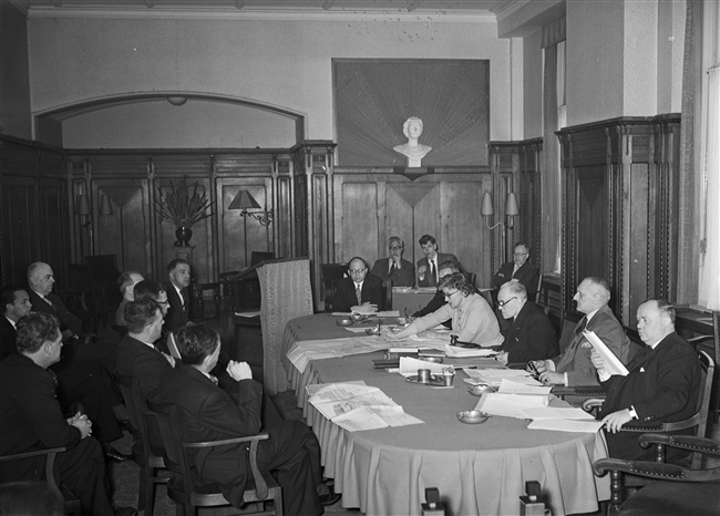Een vergadering van de Gedeputeerde Staten aan het Korte Voorhout 1. Aan tafel zitten van rechts naar links: gedeputeerde mr. H. van Riel, griffier mr. F.A. Helmstrijd, gedeputeerde dr. J.J.R. Schmal en gedeputeerde mevrouw mr. Chr. A. de Ruyter-de Zeeuw.